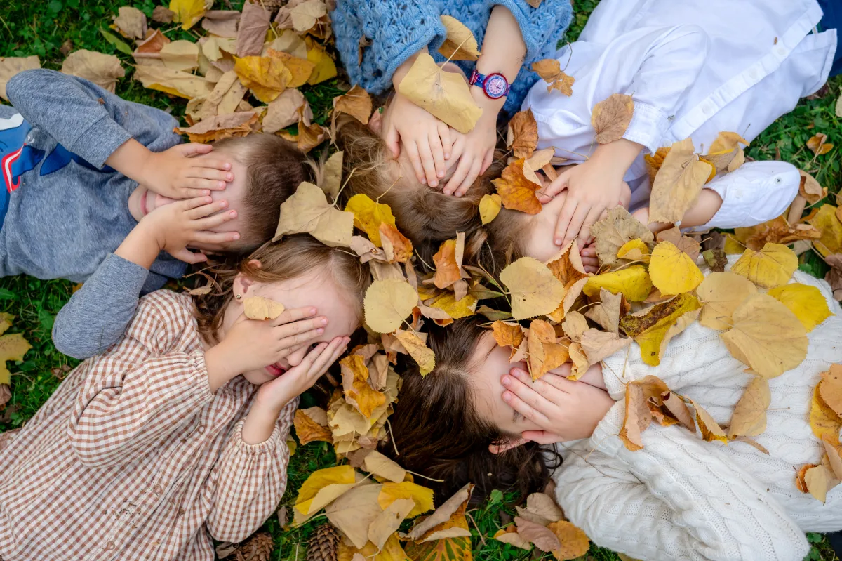 Copii jucându-se întinși în frunze de toamnă, cu mâinile la ochi — ședință foto de familie lângă Cluj-Napoca